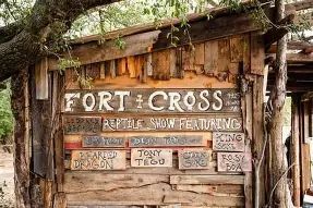 Weathered wooden shack with a hand-painted sign reading 'FORT + CROSS' and 'Reptile Show Featuring' with smaller performer plaques.