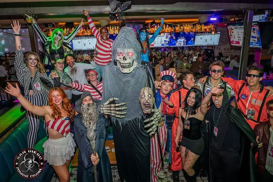 Group of people in creative Halloween costumes posing together in a bar, with a central Grim Reaper figure and others wearing striped, superhero, and patriotic outfits.