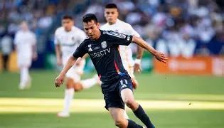 Soccer player in a dark jersey dribbling the ball on a green field with white-jersey opponents chasing in the background.