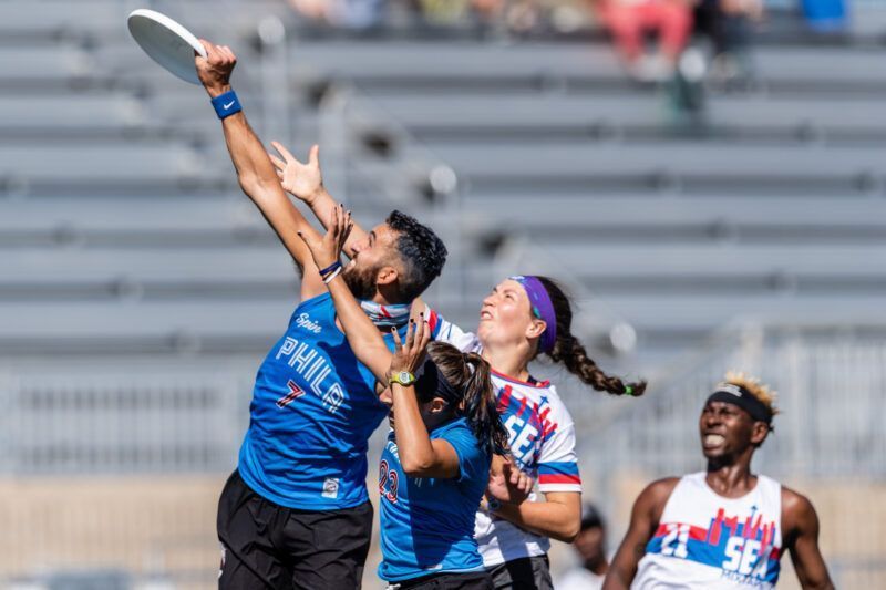 Five players in a frisbee game jump for the disc; blue jerseys battle white jersey in a sunny outdoor field, nearby spectators in bleachers.