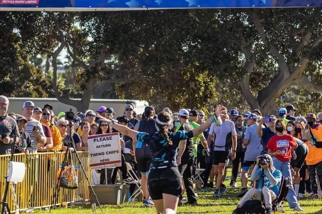 Runner with arms raised approaches the cheering crowd behind yellow barriers during a race, a 'Please return timing chips here' sign visible nearby.