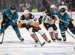 Four hockey players from two teams chase the puck on the ice, with one player in white-orange leading the chase as teal-clad opponents close in, in a crowded arena.