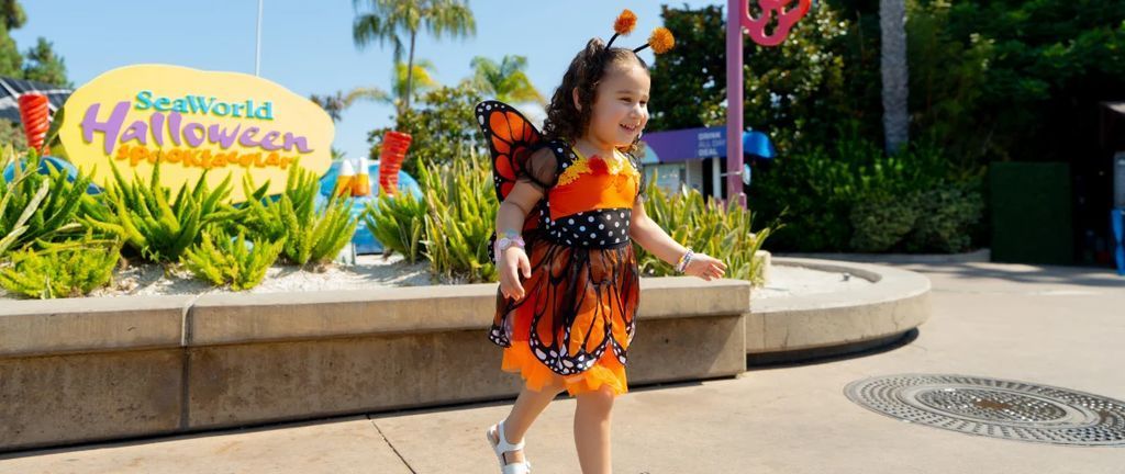 Young girl in an orange butterfly costume walking at SeaWorld Halloween Spooktacular with plants and a festive sign in the background