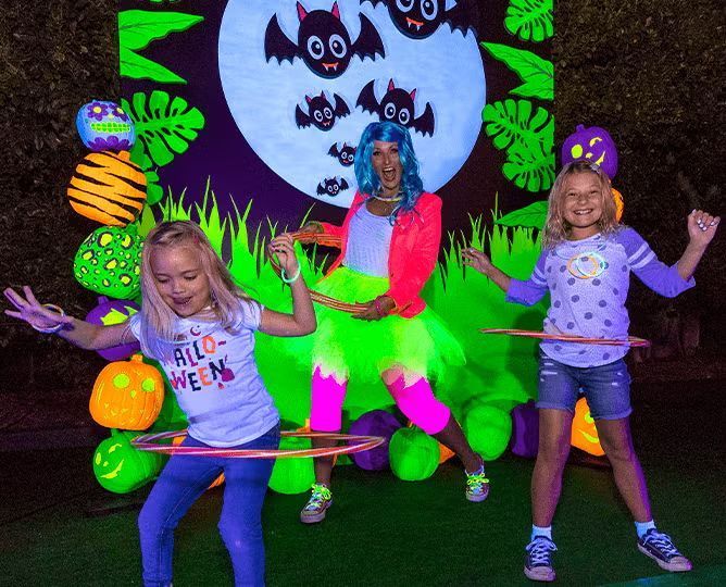 Three kids hula-hooping in a neon Halloween display with a blue moon and bats backdrop.