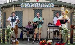 Five-member band performing on a small outdoor stage under an Oktoberfest banner, playing guitar, drums, accordion, and brass with flower arrangements in front.