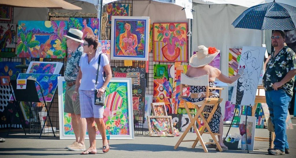 Colorful paintings at an outdoor art market with a couple walking past and a woman painting at an easel among vibrant canvases.